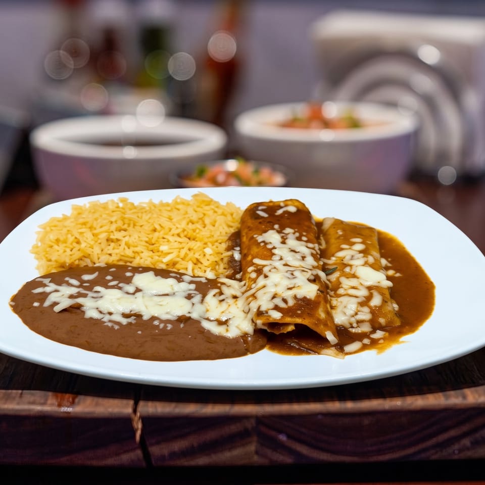 Two enchiladas with mole sauce and melted cheese, served with yellow rice and refried beans.