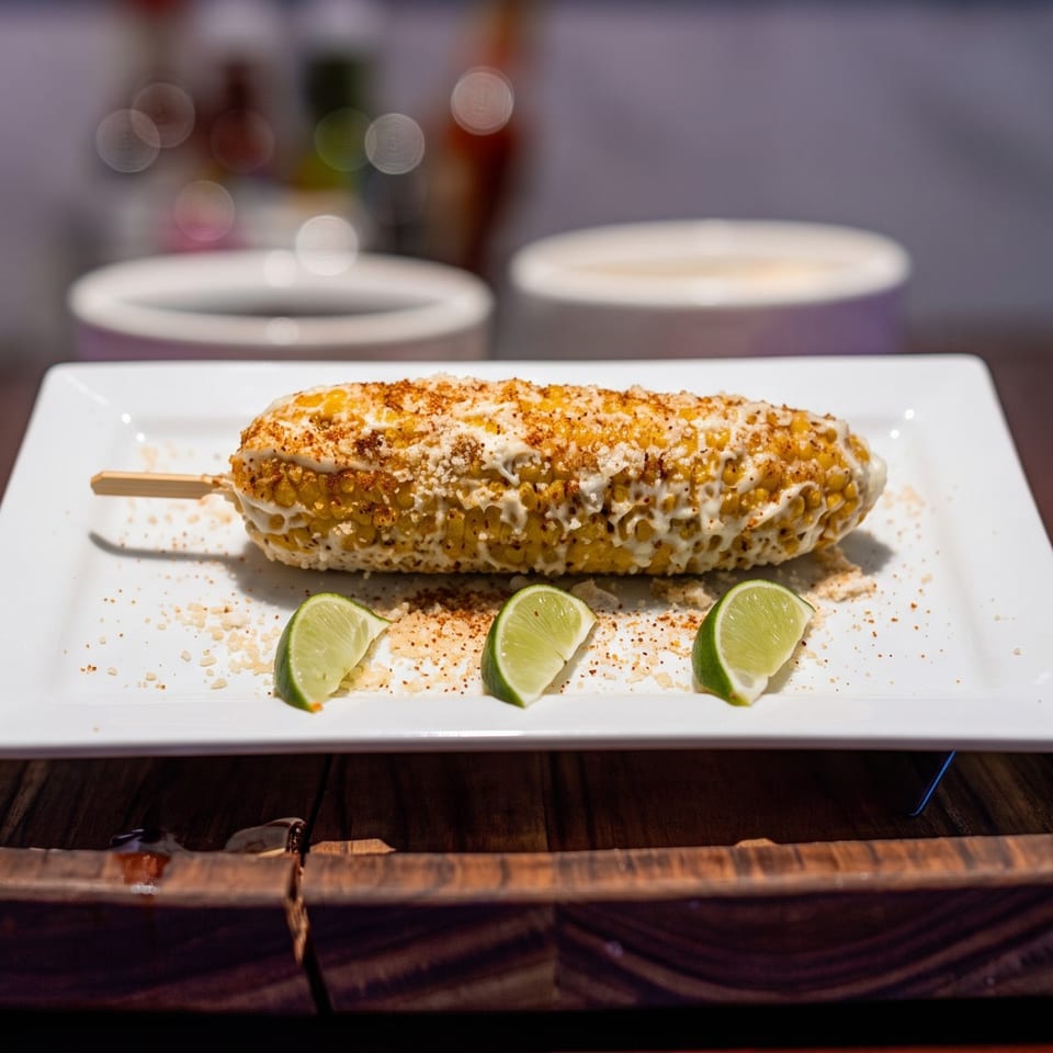 Elote, Mexican street corn, on a white plate with three lime wedges and chili powder.