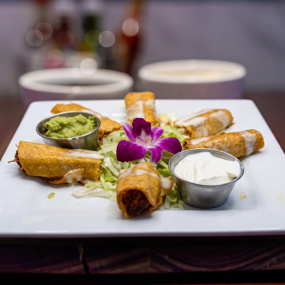 Crispy taquitos drizzled with white sauce, served with guacamole, sour cream, and a purple orchid.