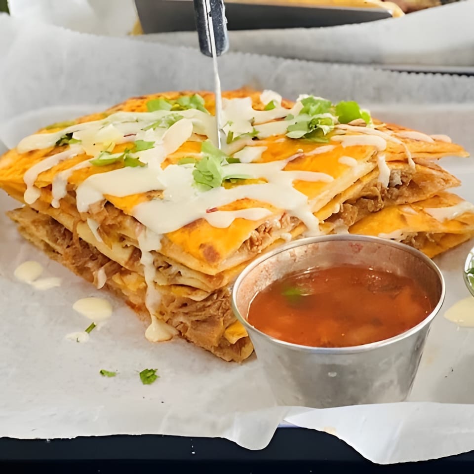 Stacked quesadilla slices drizzled with white sauce and cilantro, served with a side of salsa.